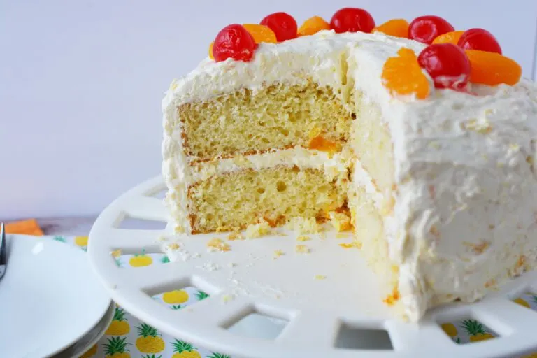 A layered cake with white frosting is topped with red cherries and orange slices. A slice has been cut out, revealing the cake's fluffy interior. The cake sits on a white cake stand with a pineapple-patterned cloth underneath.